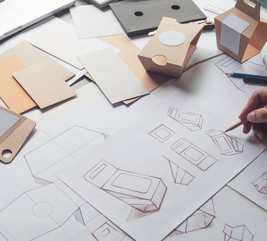 Image montrant des croquis et des prototypes de conception d'emballages sur une table, avec une personne dessinant des concepts d'emballages.